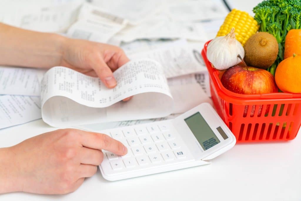 Calculating expenses beside a grocery basket and receipts, illustrating practical ways to save on groceries.