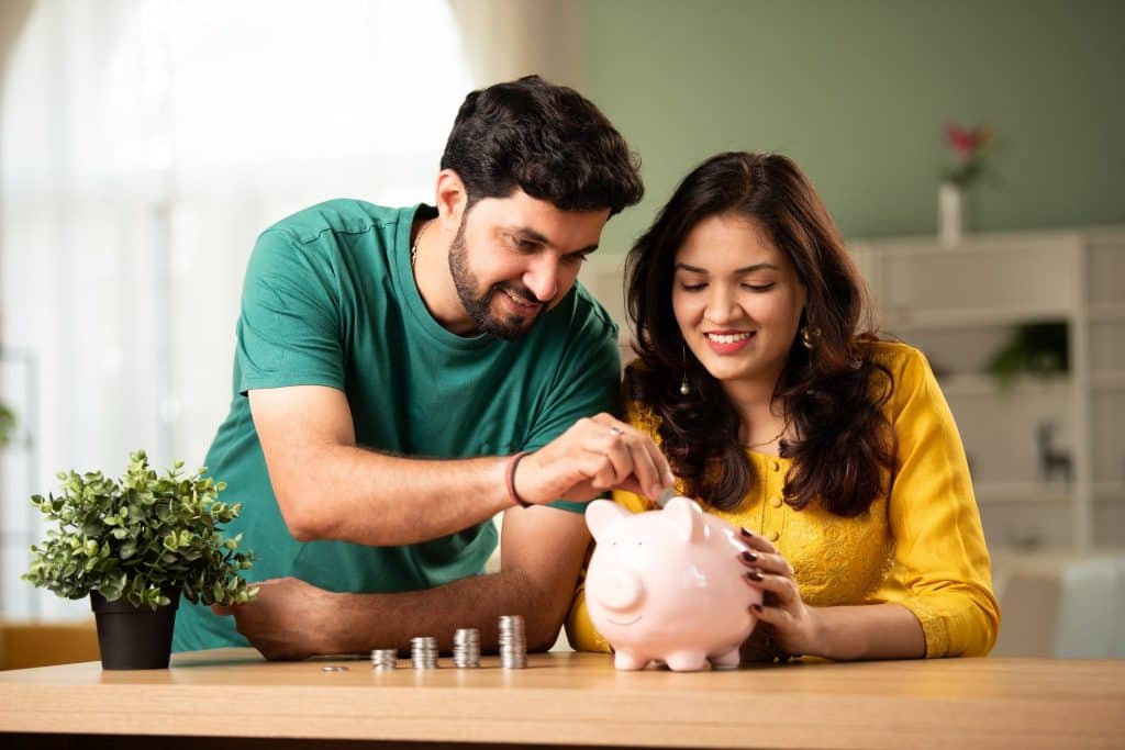 Smiling couple saving coins in a piggy bank at home, symbolizing practical tips to save money.