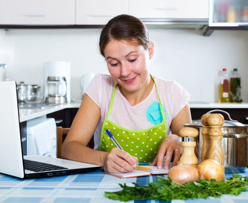 Woman planning a weekly menu at the kitchen table with a laptop and ingredients simple budget meal planning.