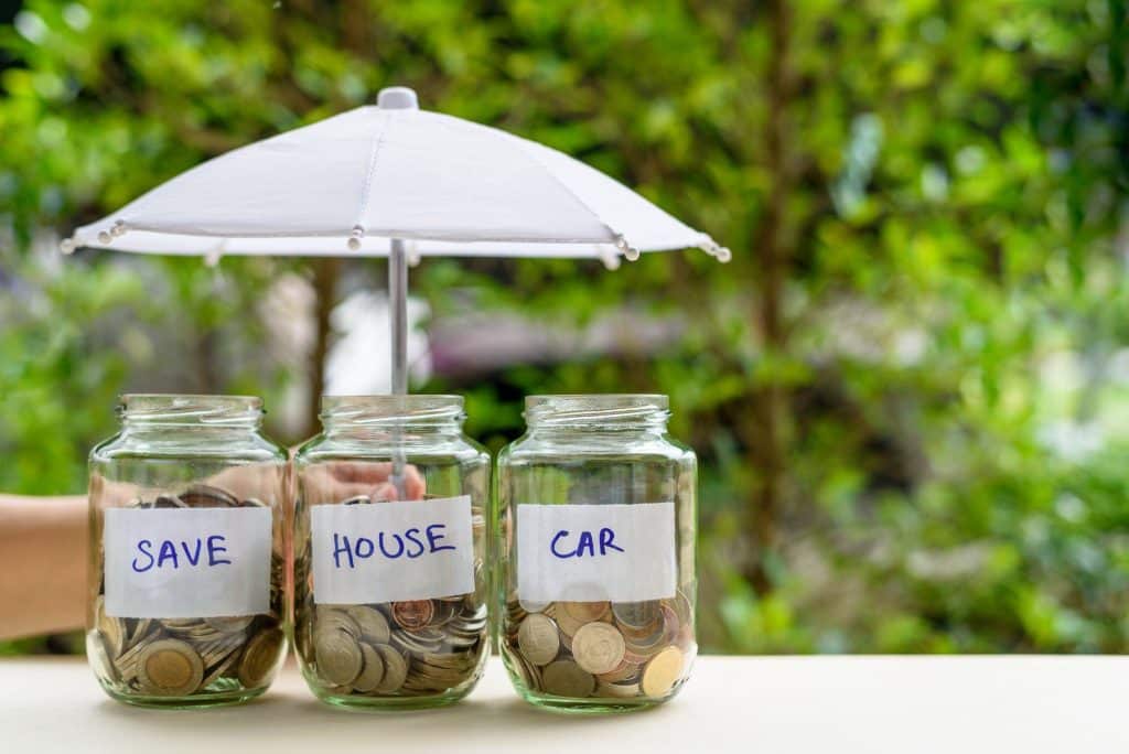 Glass jars labeled save, house, and car under an umbrella, symbolizing building emergency fund protection.