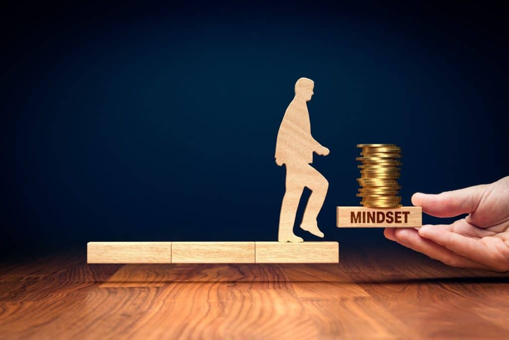 Wooden figure walking toward stacked coins labeled mindset, representing overcoming money stress and earning more with a positive outlook 