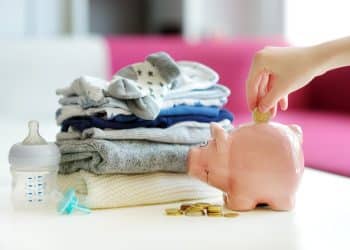 Hand placing coin in piggy bank beside baby clothes and a bottle, representing practical steps in budgeting for a baby.