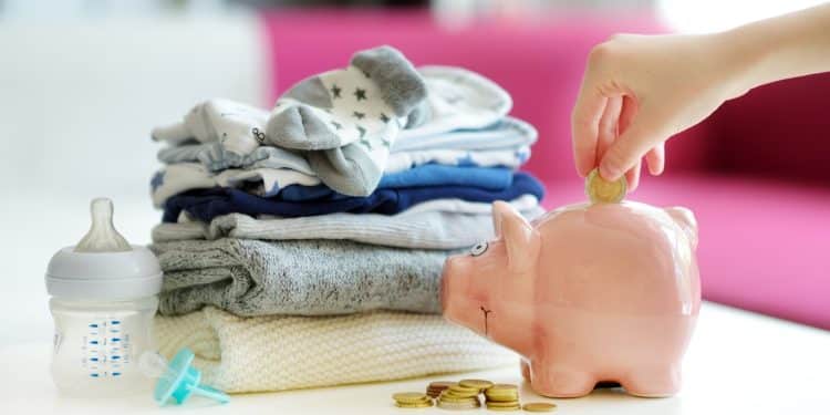 Hand placing coin in piggy bank beside baby clothes and a bottle, representing practical steps in budgeting for a baby.