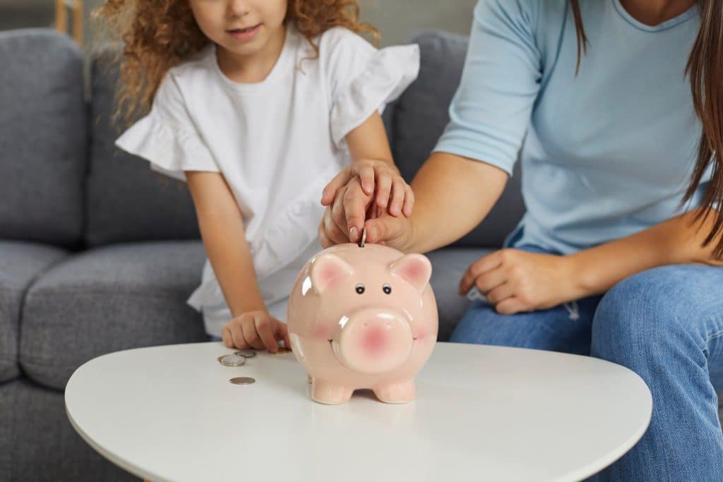 Mother teaching child the importance of budgeting for a baby by saving coins in a piggy bank.