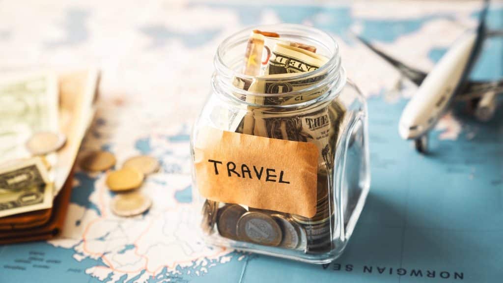 Glass jar labeled 'Travel' filled with international currency and coins on a world map, symbolizing how to travel on a budget with proper savings.