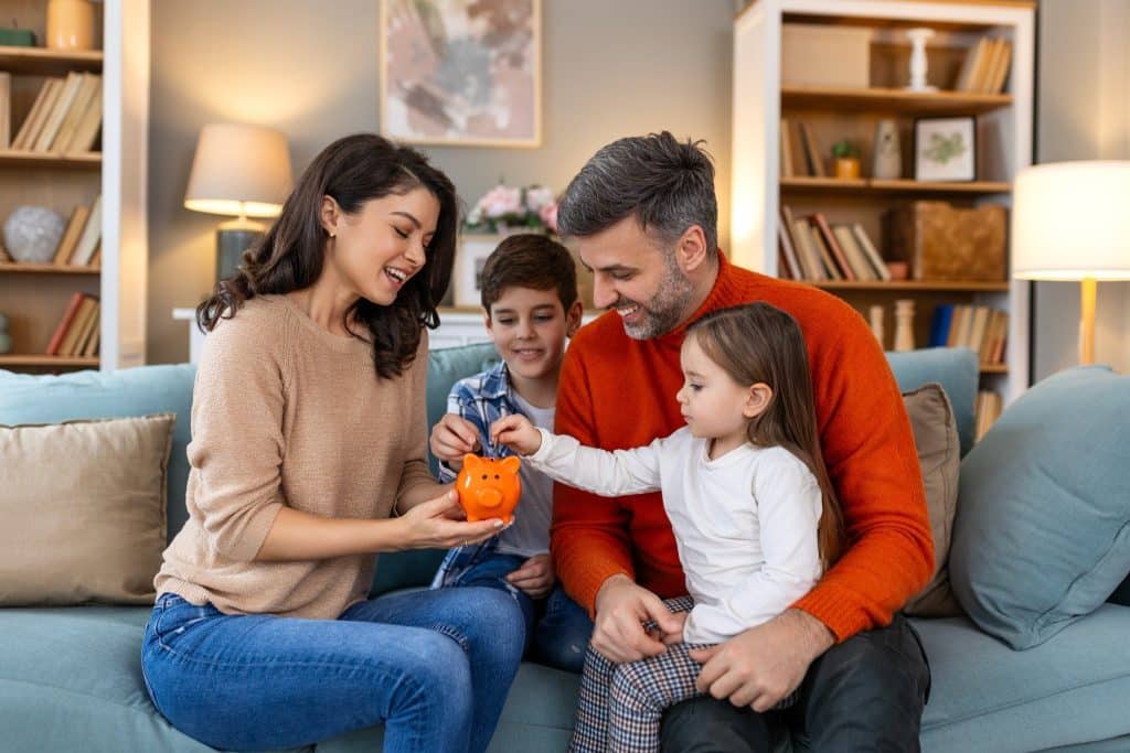 Happy family teaching kids about saving with a piggy bank, showing influence of family in breaking bad money habits early.