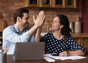 Smiling couple high-fiving at a kitchen table with laptop and papers, celebrating progress in building emergency fund goals.