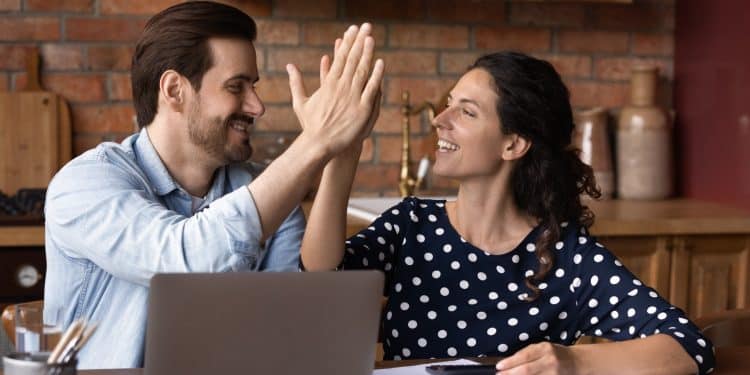 Smiling couple high-fiving at a kitchen table with laptop and papers, celebrating progress in building emergency fund goals.