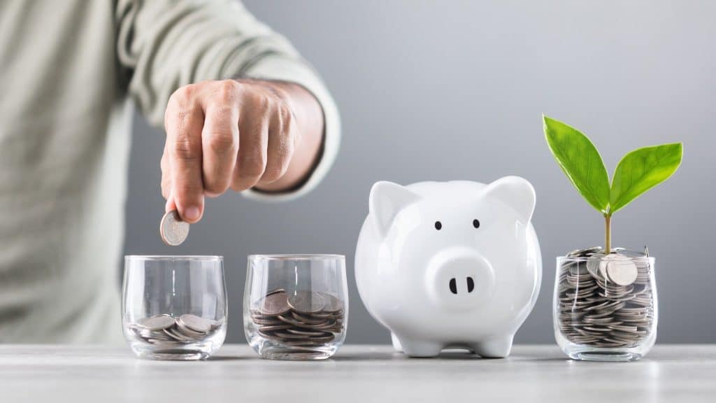 A hand placing coins into glass jars alongside a piggy bank and a growing plant, symbolizing managing monthly finances through steady savings and long-term financial growth.